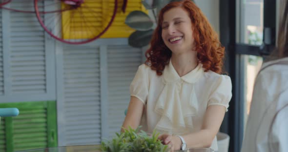 Two young business women colleagues laughing during a coffee break in a colorful boutique cafe.