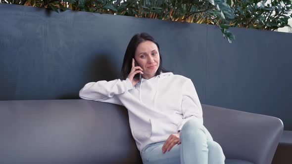 Smiling Young Business Woman of European Appearance in a White Sweater Sits in a Cozy Green Place in