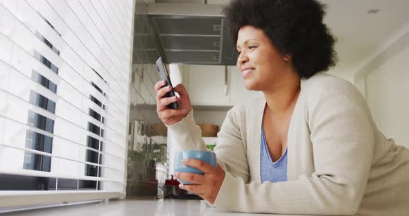 Video of happy plus size african american woman drinking coffee and using smartphone