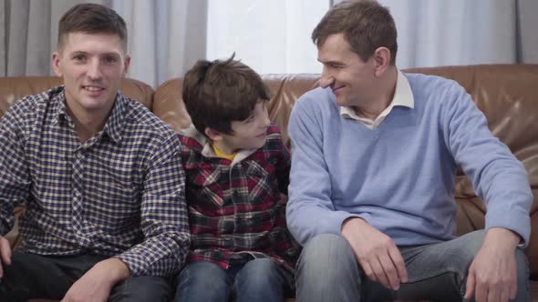 Camera Approaching To Little Caucasian Boy Sitting on Couch and Hugging His Father and Grandfather