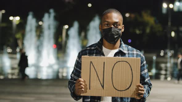 African American Man in Medical Mask Stands in Evening Night City Holding Sign Banner Cardboard Text