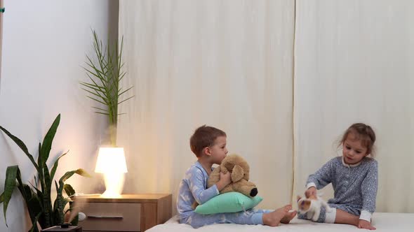 Two Smiling Preschool Toddler Children In Pajamas Playing With Teddy Bear on Bed