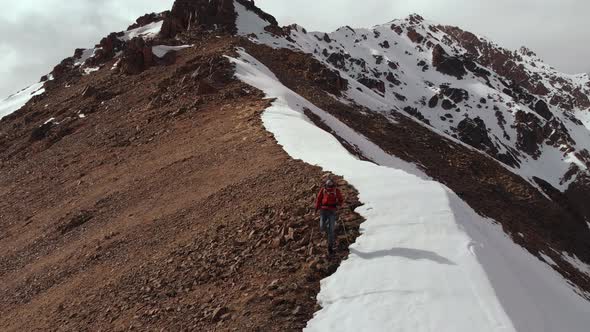 Aerial View Young Man in Sunglasses with a Backpack Goes Down the Mountain on the Crest on the