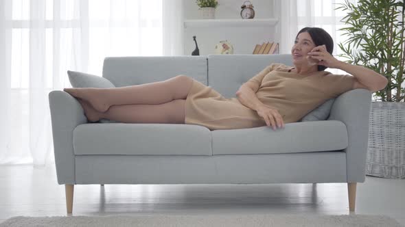 Relaxed Caucasian Senior Woman in Elegant Beige Dress Lying on Sofa and Talking on the Phone, Happy