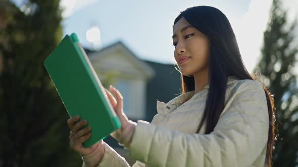 Middle Shot of Confident Smiling Asian Woman Standing in Sunrays Typing on Tablet