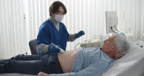 Doctor Wearing Safety Mask with Ultrasound Scan Examining Abdomen of Senior Male Patient in Clinic