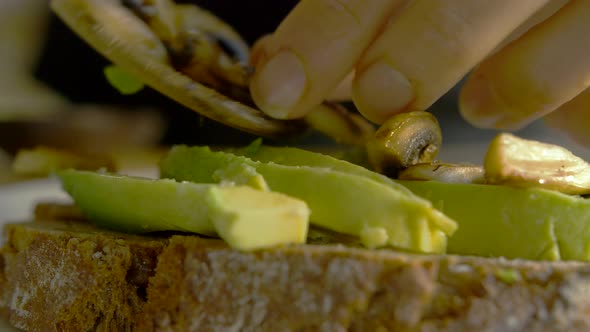 Closeup of female putting mushroom topping on avocado toast. Young white woman making healthy snack