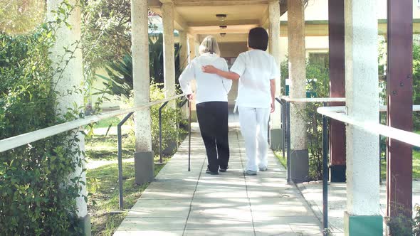 Back View of Asian Female Doctor Helps Elderly Woman with Walker in the Hospital Garden Patio Slow