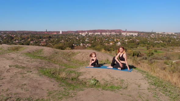 Aerial Drone View of Slender Young Mother Do Yoga Exercises with Child Daughter on High Hill By Lake