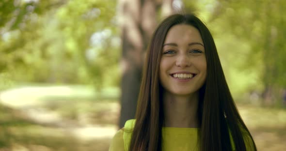 Joyful Pretty Brunette Girl Is Smiling Broadly and Playing Jokingly with Hair in Summer Day