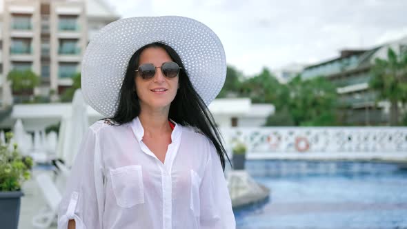 Smiling Tourist Woman in Hat and Sunglasses Walking Near Swimming Pool Admiring Luxury Resort
