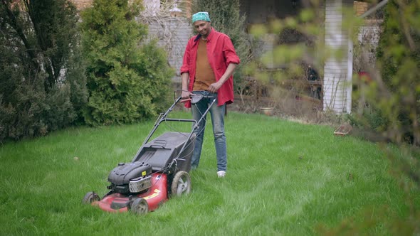 African American Young Man Scratching Head Standing on Green Backyard Lawn with Lawn Mower