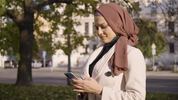 A Young Beautiful Muslim Woman Works on a Smartphone with a Smile in a Street in an Urban Area
