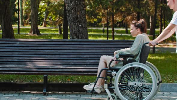 A Couple Walking in Park  a Guy Rides a Woman in a Wheelchair and About to Transfer Her on a Bench