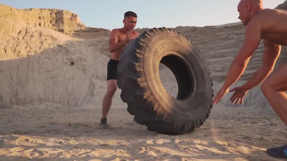 Two Athletes Train in Active Mode on the Beach Doing push-UPS and Pushing a Huge Wheel