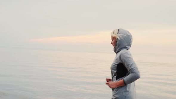 Running in Cool Weather. A Woman in a Sport Suit and a Hood Running Around the Sea or the Lake