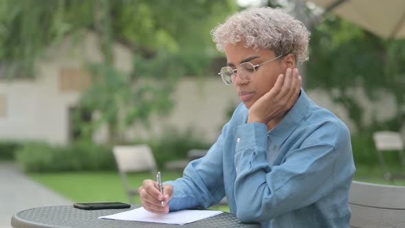 Young African Woman Thinking While Writing on Paper in Outdoor Cafe