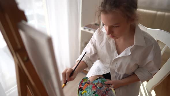 Close Up of Young Artist Sits on a Chair Near an Easel and Draws a Picture