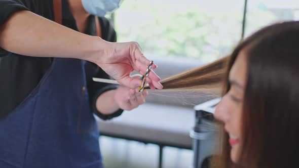 Asian male hairdresser hairstylist combing and using scissors cutting young customer's hair in salon