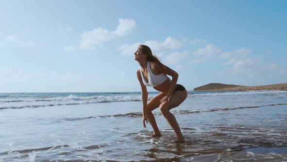 Girl in Sportswear Shorts and T-shirt Performs Jumps with Squats on the Beach Near the Ocean