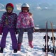 Little Girls in Outwear and Masks with Helmet Sitting on Wood Fence with Skis Near Against Mountains - VideoHive Item for Sale