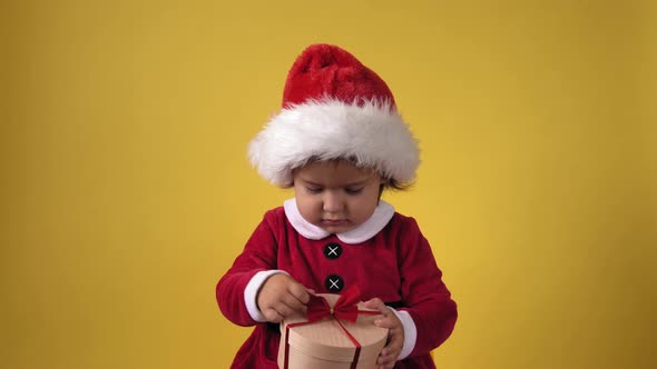 Happy Cheerful Chubby Toddler Baby Girl in Santa Suit Looking On Camera At Yellow Background