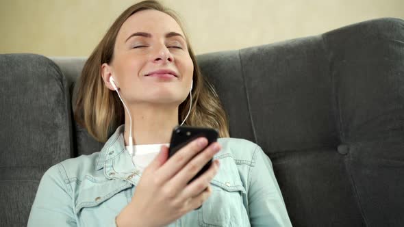 Young Woman Sitting Listening To Music with Earphones on Her Mobile Phone at Home As She Relaxes