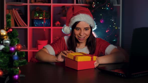 A Young Woman in a Santa Claus Costume in the Office Opens a Gift Containing Money