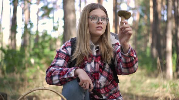 Curios Young Woman in Eyeglasses Sitting in Forest Raising Mushroom Looking at Raw Vegan Fungus in