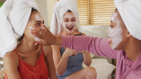 Diverse group of happy female friends with towels on heads and cleansing masks taking selfie at home