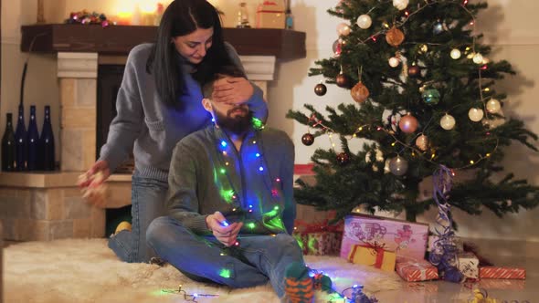Cute Joy Young Couple Sitting at the Christmas Tree on the Floor in the Room. The Woman Giving Gift