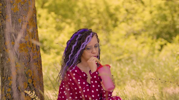 Young Woman in Glasses Red Dress Drinking Refreshing Beverage Sitting Near Tree in Sunny Windy