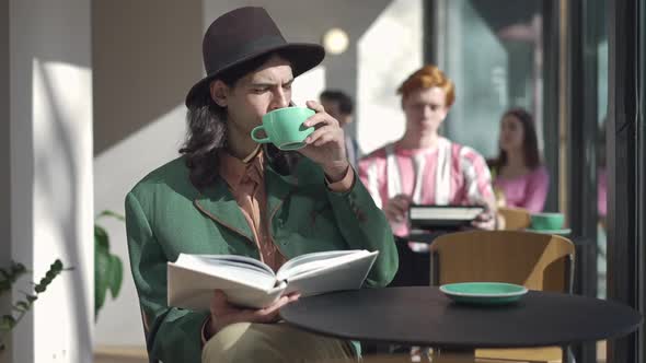 Intelligent Stylish Man Drinking Coffee Reading Book in Vintage Cafe Indoors on Sunny Day