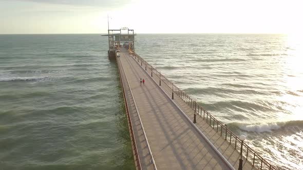 Aerial view of teenagers boy and girl walk by hand on pier in sea foaming waves love on beach summer