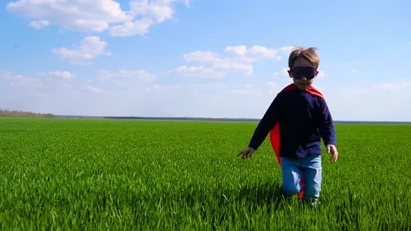 Joyful Child in a Superhero Costume, a Red Cloak and a Mask Runs Through the Green Grass, Looking at