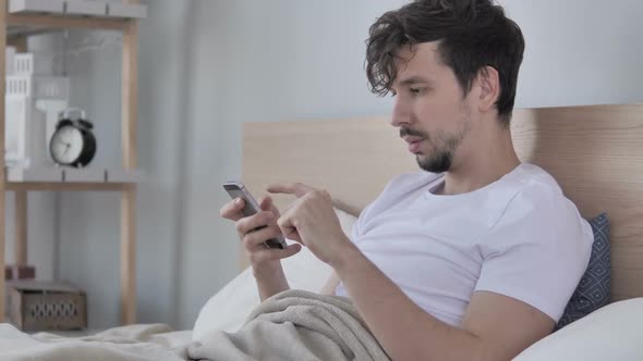 Casual Young Man Celebrating Success While Using Smartphone in Bed