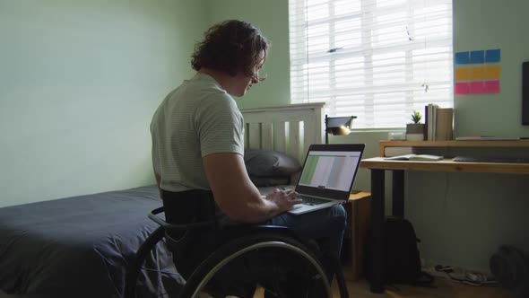 Focused caucasian disabled man in wheelchair using laptop in bedroom