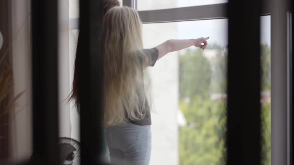 Two Little Girls with Blond and Dark Hair Looking Out the Window in the Living Room. Brunette Girl