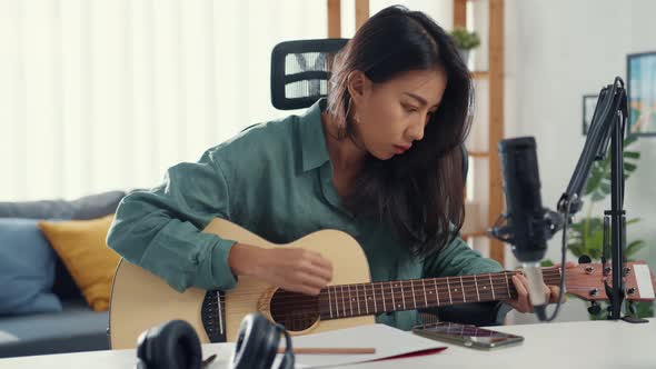 Happy asia woman songwriter play acoustic guitar listen song from smartphone think and write notes.