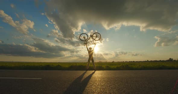 The Cyclist is Standing on the Edge of the Road and Lifting the Bike Above Him