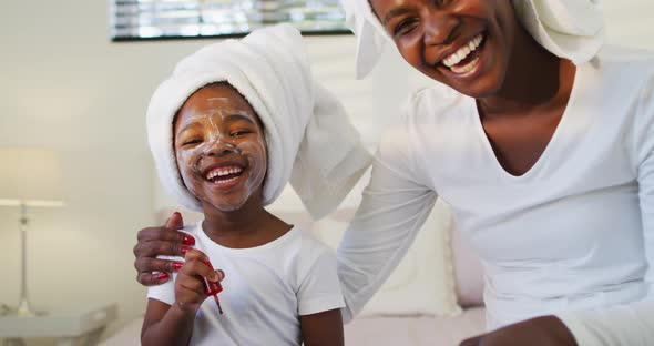 Happy african american mother and daughter wearing towels sitting on bed and looking to camera