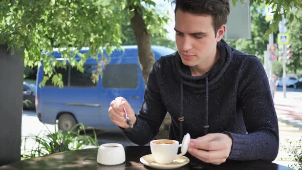 Young Man Mixing Sugar and Drinking Coffee Sitting in Cafe Terrace