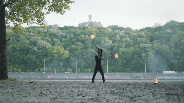Young Slim Man in Black Clothes and Mask Performing a Show with Flame Standing on Riverbank
