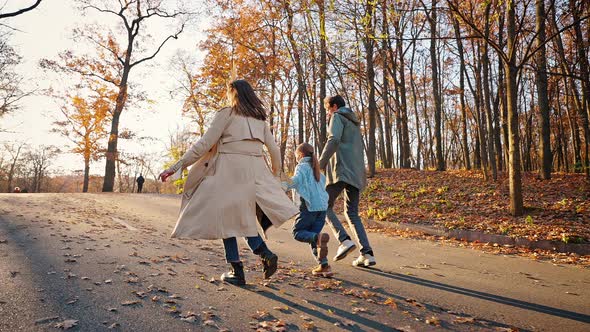 Young Parents Running By Paved Road in Autumn Park with Little Daughter Jumping Hanging on Their