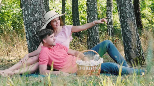 Picnic Time. Happy Young Couple Sitting and Smiling Outdoors. Girl Pointing Her Hand with Finger