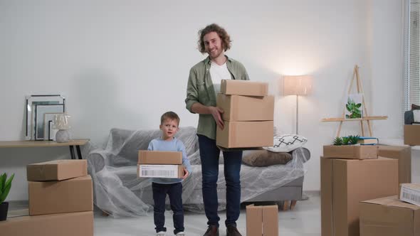 Young Handsome Father and Son Stand with Cardboard Boxes in New Apartment with Furniture Covered