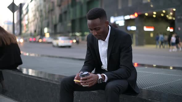 Young guy drinking coffee from a disposable cup after work and texting on phone