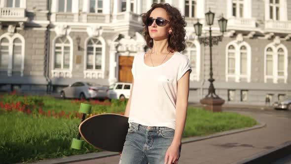 Beautiful Young Woman in Sunglasses Walking in the Old City Street Holding Her Longboard in the