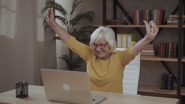 Cheerful Older Woman in Smart Casual Wear and Eyeglasses Is Looking at Laptop Screen and Holding