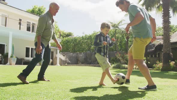 Happy caucasian grandfather with adult son and grandson playing football in garden